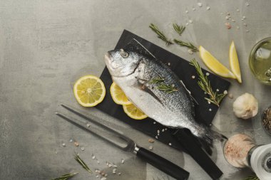 Limonlu taze dorado balığı, biberiye ve siyah kesme tahtasındaki baharatlar, yakın çekim. Gri masadaki malzemeli çiğ deniz ürünleri. Sağlıklı gıda konsepti
