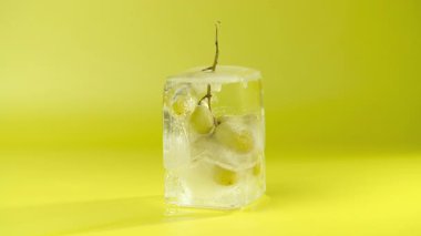 Buz küpündeki üzümler, yakın plan. Buzlar erir ve bir demet yeşil üzüm ortaya çıkar. Donmuş meyve. Sarı arkaplan.