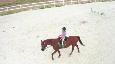 Genç bir binici, kumlu bir arenada kestane renkli bir ata biniyor ve binicilik sporunun neşesini sergiliyor.