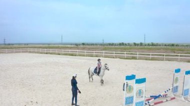 Bir çocuk kumlu bir arenada, bir eğitmenin rehberliğinde küçük gri bir at üzerinde binicilik becerileri geliştiriyor.