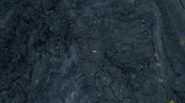 Bir kadın katılaşmış siyah lavların üzerinde, parlak elbiseleriyle karanlık ve engebeli arazisi arasındaki katı zıtlığı sergiliyor..