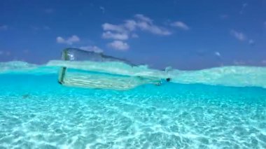 Maldivler 'deki bir sualtı sahnesinde cam bir şişe kristal berrak sularda yüzüyor, kumlu okyanus tabanı ve yukarıda parlak bir gökyüzü ile çevrili..