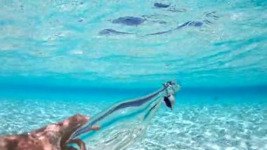 Bir el Maldivler 'de su altında uzanır, cam bir şişeyi kavrar, etrafını turkuaz su sarar ve yukarıda mavi bir gökyüzü vardır..