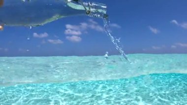 Bir el, arkasında mavi bir gökyüzü olan Maldivler 'in berrak sularına su döken bir cam şişeyi tutar..