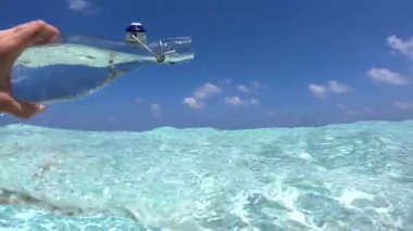 Bir el açık bir cam şişeyi tutar, berrak sulara su döker, Bora Bora 'da su altında yakalanır, arka planda bir plaj vardır..