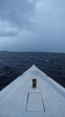 Bir tekne Maldivler 'in dalgalı sularında, bulutlu gökyüzünün altında, uzak bir kıyı ve iletişim kulesiyle yol alır..