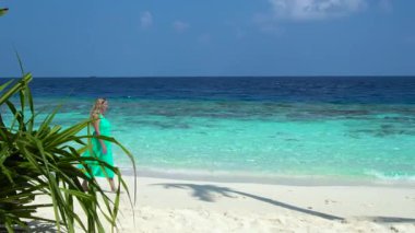 Kadın Maldivler 'de kumlu bir sahil boyunca yürüyor, berrak turkuaz suların yanında ve mavi gökyüzünün altında..