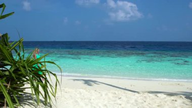 Kadın Maldivler 'de kumlu bir sahilde duruyor, sığ turkuvaz suları ve yumuşak dalgalarıyla. Yeşil yapraklar sahneyi çerçeveliyor.