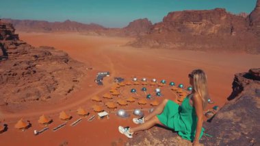 Yeşil elbiseli bir kadın, baloncuklu çadırlar ve geleneksel çadırları olan, Ürdün 'deki Wadi Rum, Wadi Çölü' ndeki eşsiz bir çöl kampına bakan bir kayanın üzerinde oturuyor..