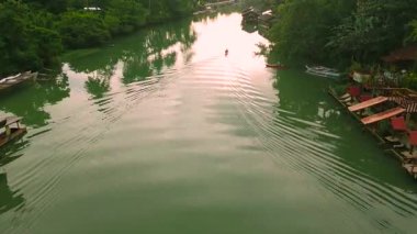 Amazon Nehri kıyısında sık ağaçlar ve geleneksel evlerle çevrili küçük bir teknede kürek çeken bir adamın bulunduğu yeşil bir nehrin havadan görünüşü..