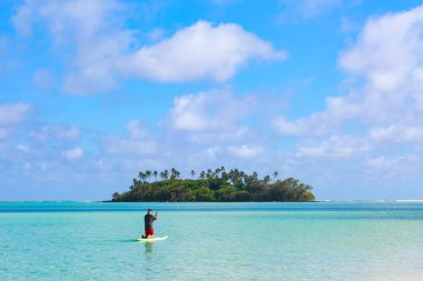 Güzel bir turkuaz gölün ortasındaki tropik bir adaya doğru kürek çeken bir adam. Muri, Rarotonga, Cook Adaları 'nda çekilmiş, Taakoka Adası' na doğru bakıyor.