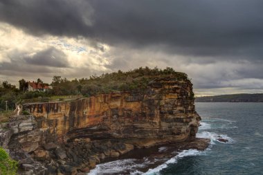 Sydney Harbour, Sydney, Avustralya 'nın girişindeki uçurum. Güneş ışığı yukarıdaki kasvetli bulutların arasından doğuyor.