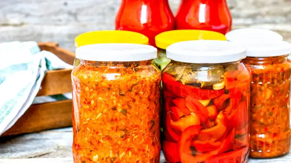 Glass jars with pickled red bell peppers and pickled cucumbers (pickles) isolated. Jars with variety of pickled vegetables, jars with zacusca and bottles with tomatoes sauce. Preserved food concept.