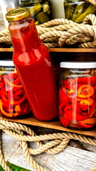 Wooden crate with bottles with tomatoes sauce and glass jars with pickled red bell peppers isolated in a rustic composition. Jars with variety of pickled vegetables preserved food concept.