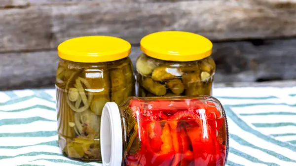 Glass jars with pickled red bell peppers and pickled cucumbers (pickles) isolated. Jars with variety of pickled vegetables. Preserved food concept in a rustic composition.