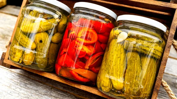 Glass jars with pickled red bell peppers and pickled cucumbers (pickles) isolated in wooden crate. Jars with variety of pickled vegetables. Preserved food concept in a rustic composition.