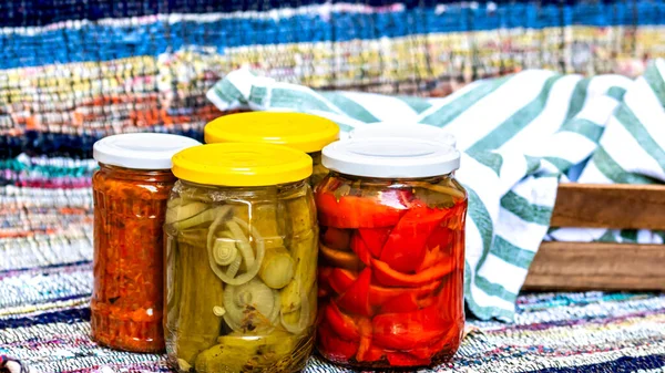 Glass jars with pickled red bell peppers and pickled cucumbers (pickles) isolated. Jars with variety of pickled and canned vegetables. Preserved food concept in a rustic composition.