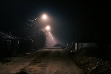 Karanlık ve sisli bir kış gecesinde, uzak mesafede parlayan sokak ışıklarıyla boş, kasvetli bir sokak.