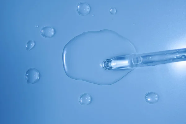 Water bubbles with cosmetic liquid drops of serum on a blue background of a laboratory glass pipette. Close-up of a pipette with drops.