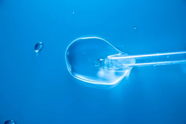 Water bubbles with cosmetic liquid drops of serum on a blue background of a laboratory glass pipette. Close-up of a pipette with drops.