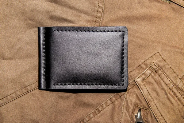 Black handmade leather wallet on brown tactical pants. Male background. View from above.