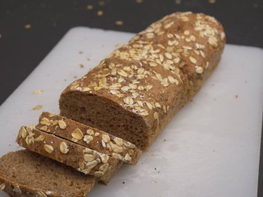 Kısmen dilimlenmiş taze organik kabuklu ekmek beyaz bir kesme tahtasının üzerinde yatıyor. Kırıntılar ve yulaf ezmeleri ekmeğin etrafına saçılmış..