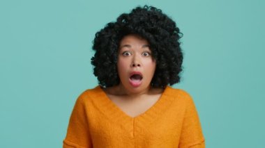 Şok olmuş, ağzı açık, hayret ve hayretle yüzüne dokunan Afro-Amerikan kadın portresi. Kıvırcık saçlı model yavaş çekim stüdyosu mavi arka planda 4K
