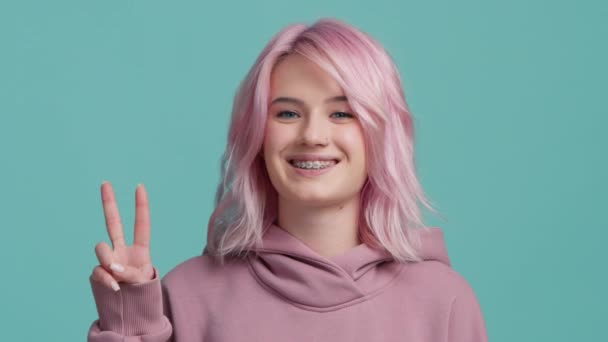 満面の笑みピンク色の髪をした陽気な女の子ジェスチャー Sign Smiling Camera Posing Teal Blue Studio背景 勝利ジェスチャーを示すカジュアルファッショナブルな女性 正の動機付けの概念 — ストック動画