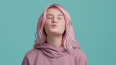 Nazik pastel rengi kapüşonlu pembe saçlı güzel bir kadın açık mavi stüdyo arka planında hava öpücüğü gönderiyor, yavaş çekim arkadaşlığı, aşk, flört ve romantik konsept görüntüler.