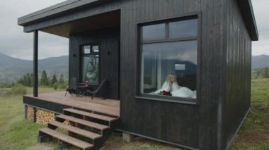 Kadın, beyaz battaniyeye sarılmış, sabah kahvesini yudumluyor. Kadın turist, pencereden göründüğü gibi sinematik doğal manzaranın tadını çıkarıyor. Yüksek kalite 4k görüntü