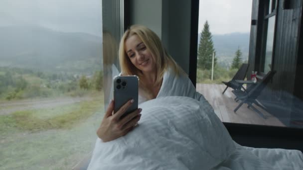 美丽的 高加索女孩采取自拍的早晨 女人在一个舒适的房间里享受早晨 房间里有全景窗户和周围的山水 高质量的4K镜头 — 图库视频影像