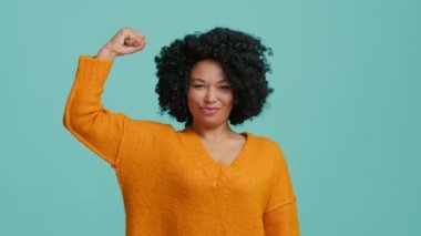 Kendinden emin Afro-Amerikan kadın bunu yapabileceğini gösteriyor ve kollarında kaslarını gösteriyor. Güçlü kız, mavi arka planda izole edilmiş spor başarılarıyla gurur duyuyor. Kız gücü kavramı