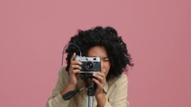 Odaklanmış Afrika kökenli Amerikalı kadın fotoğrafçının belden yukarı portresi, analog kamerayla tripoda poz veriyor, izole edilmiş arka planda parlak pembe fotoğraf stüdyosunda duruyor, kameraya ve vizörüne bakıyor.