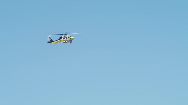 澄んだ青い空を飛んで白い黄色のヘリコプターのスローモーションショット ロサンゼルス郊外での野火を区別した後 青い空を飛んで消防ヘリコプター シネマティックローアングルショットヘリ — ストック動画