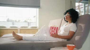 Yavaş çekim, okuma gözlüklü güzel Afrikalı Amerikalı kadın oturma odasında elinde patlamış mısırla kanepede uzanıyor uzaktan kumandayla televizyonda ilginç bir film izliyor. 4K içinde hafta sonu