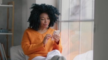 Rahat Afro-Amerikan kadın cep telefonuyla sosyal medyayı izliyor, soğuk kış günü pencerede oturuyor. Akıllı telefon teknolojisi kullanan güzel kıvırcık saçlı kadın internette yavaş çekimde satış yapıyor.