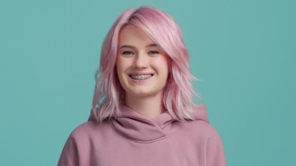 スタイリッシュなピンク色の髪型とカメラに積極的にまっすぐ見て歯のブラケットを持つ若い20代の女性学生のスローモーションの肖像画と公然と笑みを浮かべて オンラインインタビューや教育の概念 — ストック動画