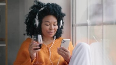 Genç gülümseyen kıvırcık saçlı, kırmızı şarap içip dans eden, penceredeki kablosuz kulaklıkla müzik dinleyen Afro-Amerikan kadın. Kadın eğlencesini bir bardak şarapla kapat.