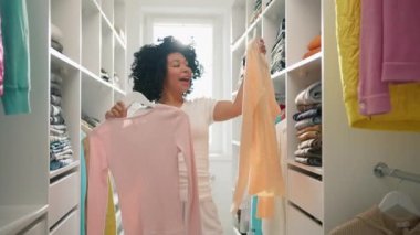 Renkli dans eden ve şarkı söyleyen tatlı, komik bir kadın. Randevuya ya da geceye hazırlanırken. Ticari yavaş çekim. Güzel bir Afrikalı genç Amerikalı kadın iş için kıyafetlerini evde seçiyor.