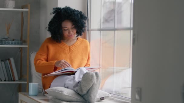家の窓辺に座って本を読んで魅力的な若い女性 スタイリッシュなインテリアのリビングルームの窓 穏やかなアフリカ人女性でオレンジセーター読書本で座っている間に窓室内 — ストック動画