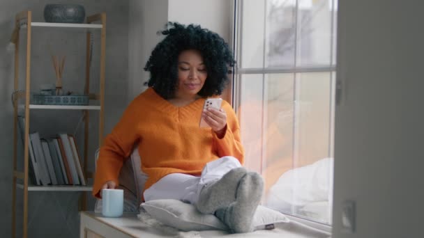 快乐的非洲裔美国人模式使用智能手机装置在家里 拥有一头卷曲黑发的女性博主在手机上订阅新的社交媒体 购买互联网 在线订购应用程序产品 — 图库视频影像