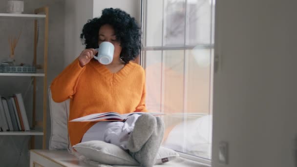 Spokojna Afrykanka Pomarańczowym Swetrze Czyta Książkę Siedząc Przy Oknie Środku — Wideo stockowe