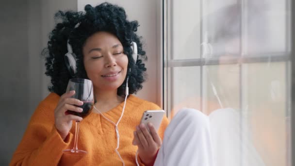 用一杯酒来招待女性 年轻的非洲裔美国女人 满脸笑容 卷曲的头发 一边喝着红酒 一边跳舞 一边用无线耳机在窗前听音乐 — 图库视频影像