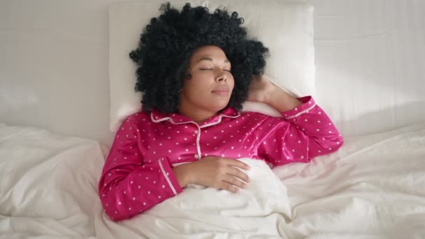 ピンクのパジャマを着たかなり新鮮なアフリカ系アメリカ人女性が快適なベッドの上に横たわっている肖像画のビューをトップダウン 寝室で朝に寝ている巻き毛の髪の色の美しい女性 健康と休息の概念 — ストック動画