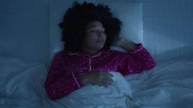 Genç, güzel, gülümseyen Afro-Amerikan bir kadının yatak odasında rahat bir şekilde uyuduğu bir manzara. Gece mavi renkleri ve soğuk zayıf sokak lambası ışığı pencere 4K 'de parlıyor.