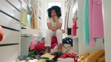 4K numaralı dolapta kıvırcık saçlı tatlı Afro-Amerikan kadın. Gardırobun ortasında oturan, kıyafetleri dağıtan, renkli, komik, üzgün kadının ağır çekimleri. Ne giyeceğini bilmiyor.