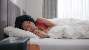 Hoş renkli güzel bir kadın yatak odasında 4K yastığında mutlu görünüyor. Genç, mutlu, Afro-Amerikan bir kadının portresi. Kameranın önünde gülümsüyor. Sabah uyandıktan sonra yatakta yatıyor.