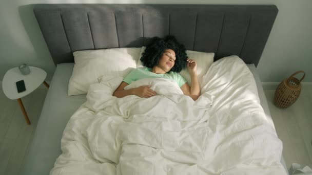 若いアフリカ系アメリカ人の女性がベッドに横たわって 明るい太陽の光を見て彼女の目を開けて寝室を照らした 色の美しい眠っている女性 ベッドルーム 健康と休息のコンセプトでトップダウンの朝 — ストック動画
