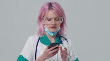 Koruyucu gözlük takan çekici genç bayan doktor cep telefonuyla hastaya danışmanlık yapıyor. Tıbbi terapist asistanı konferansında. Portre görüntüsü. Teletıp salgını konsepti 4K