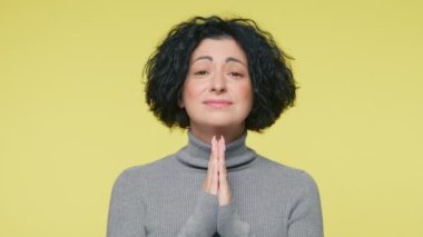 40 'lı yaşlarda komik, olgun bir kadın gri balıkçı yaka kazak giyer, dua ederken el ele tutuşur, dilek tutar, sarı stüdyo arka planında parmaklarını çapraz tutarsın.